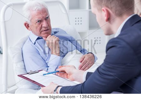 Senior man in a hospital bed about to sign his last will in the presence of the lawyer
