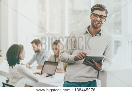 Young and successful. Happy young man holding digital tablet and looking at camera while his colleagues working in the background