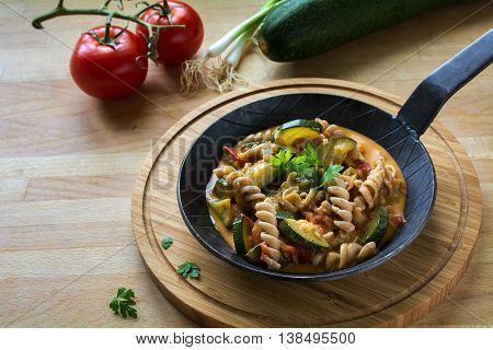 Vegetables with whole grain pasta in a rustic pan on a wooden kitchen board healthy vegetarian meal to lose weight with zucchini; tomatoes and spring onions copy space selected focus narrow depth of field