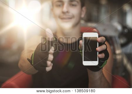 sport, bodybuilding, lifestyle, technology and people concept - happy young man with smartphone showing thumbs up in gym