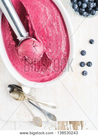 Homemade bluberry ice cream and scooper in mold served with fresh berries, silver spoons on white rusric wooden background, top view