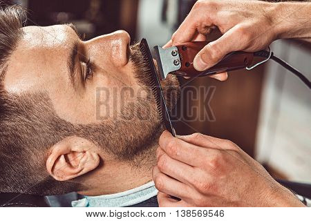 Hipster client visiting barber shop. The hands of young barber making the cut of beard