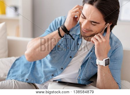 Enjoy the sounding. Cheerful delighted bearded man using tablet and resting on the couch while listening to music