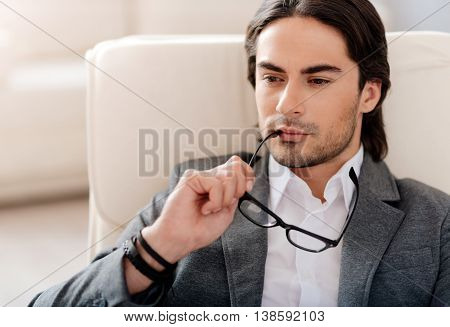 Mull it over. Concentrated handsome man sitting in the chair and holding his glasses while being involved in thoughts