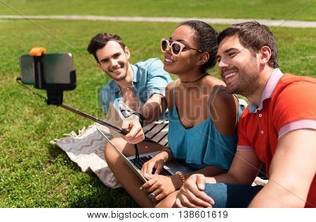Cheese. Happy and merry youth making selfie photo using mobile phone and selfie stick while sitting on the grass in the park and communicating