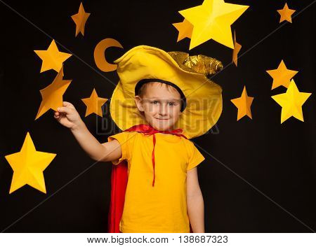 Five years old boy in stargazer costume, standing among paper stars and moon, against black background