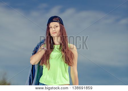 Teenager girl in cap listening to music with earphones outdoor. Portrait of young woman against sky.