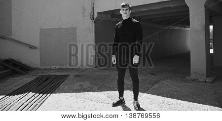 Black-and-white photo of an athlete men in a black slinky dress against the backdrop of the concrete bridge and wall