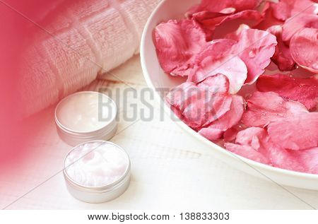 Pink rose petals in bowl, cosmetic moisturizer emollient in containers, white towel, pink natural blur, creamy tones