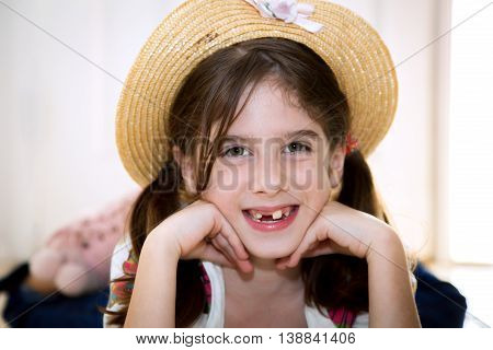 A cute young snaggletooth girl smiles with her chin on her hands. She is wearing a straw hat and laying on her stomach.