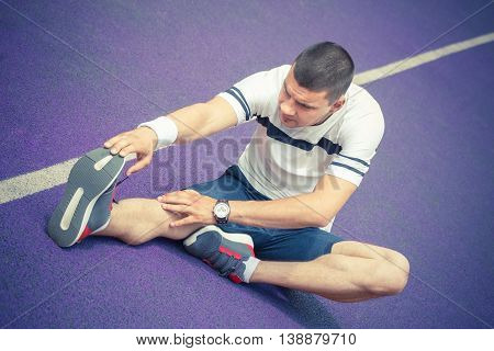 Man stretching hamstring leg muscle on the purple running track while preparing for morning workout. Caucasian sport fitness model outdoors. Recreation and healthy lifestyle concepts.