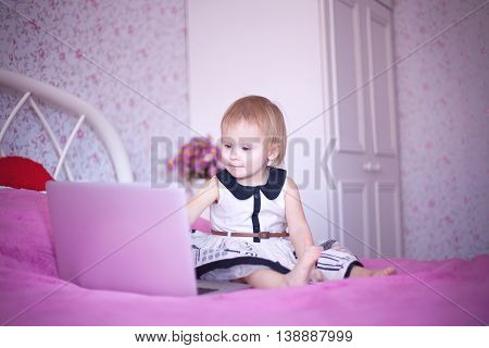 baby in pink room sitting on the bed and playing or watching cartoons price. Girl sitting at home. little princess in dress uses advanced fashion technology