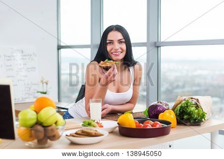 Close-up portrait of smiling woman eating diet vegetarian sandwich with vegetables for breakfast in morning, looking at camera