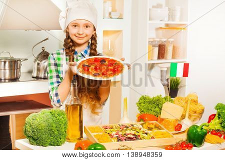 Cute young Italian girl in cook's uniform holding plate with tasty pizza in the kitchen full of kitchen-stuff