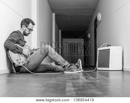 Photo of a man sitting playing his electric guitar in front of a small combo amplifier in a hallway. Black and white version.
