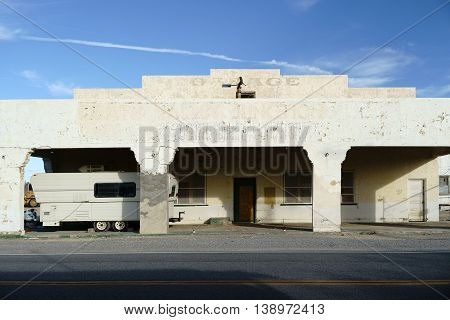 A camping trailer parked under the canopy of a rundown garage with a yellowed facade.