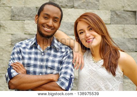 Interracial charming couple wearing casual clothes posing for camera and embracing in front of grey brick wall.