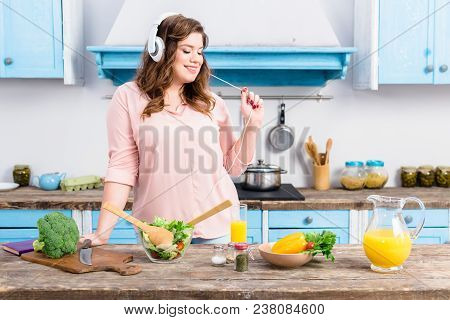 Cheerful Overweight Woman Listening Music In Headphones At Table With Fresh Vegetables In Kitchen At