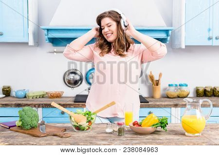 Cheerful Overweight Woman Listening Music In Headphones At Table With Fresh Vegetables In Kitchen At