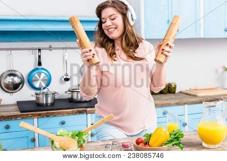 Cheerful Overweight Woman In Headphones With Wooden Salt And Pepper Grinders In Hands In Kitchen