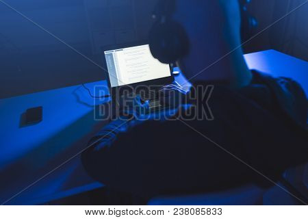 cybercrime, hacking and technology concept - male hacker with headphones and coding on laptop computer screen wiretapping or using computer virus program for cyber attack in dark room