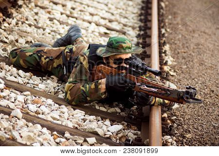 The Soldier Or Hunter In Military Uniform Is Lying Down On Railroad Track And Aiming With Crossbow W