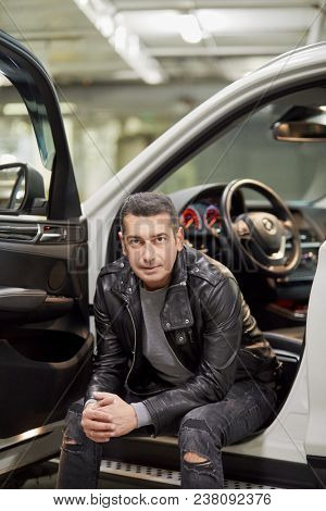 Man in jeans and leather jacket sits on doorsill of modern white car at underground parking.