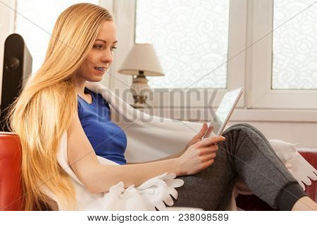 Young Woman Lying On Sofa Under Warm Blanket, Relaxing Using Tablet, Reading News And Checking Socia
