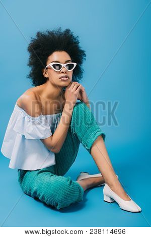 Beautiful Young Woman In Stylish Off-the-shoulder Top And Sunglasses Sitting On Floor On Blue