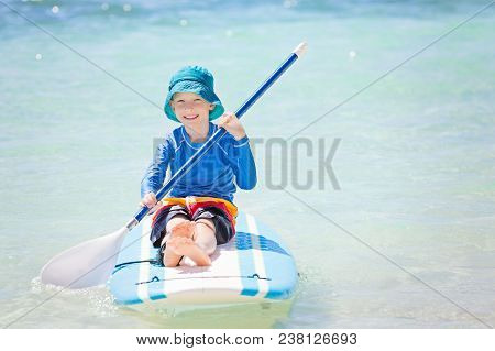 Smiling Little Boy In Rashguard Holding Paddle Sitting At Surf Board Enjoying Tropical Vacation