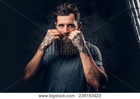 Brutal Bearded Male With Tattoos On Arms Touches His Moustache Over Grey Background.