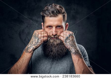Brutal Bearded Male With Tattoos On Arms Touches His Moustache Over Grey Background.