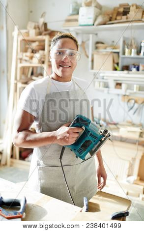 Female carpenter with electric fretsaw looking at camera through protective eyeglasses in workroom