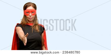 Middle age super hero woman wearing red mask and cape holds hands welcoming in handshake pose, expressing trust and success concept, greeting isolated blue background