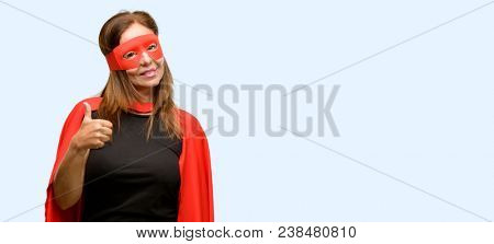 Middle age super hero woman wearing red mask and cape smiling broadly showing thumbs up gesture to camera, expression of like and approval isolated blue background