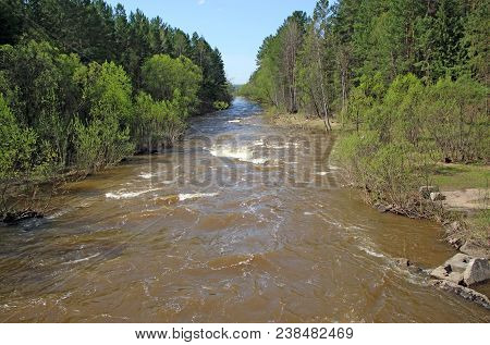 Landscape Image Of Small And Fast River Carries Its Water Among Green Trees And Bushes, And Blue Clo