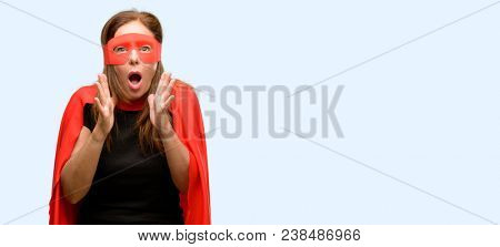 Middle age super hero woman wearing red mask and cape stressful keeping hands on head, terrified in panic, shouting isolated blue background