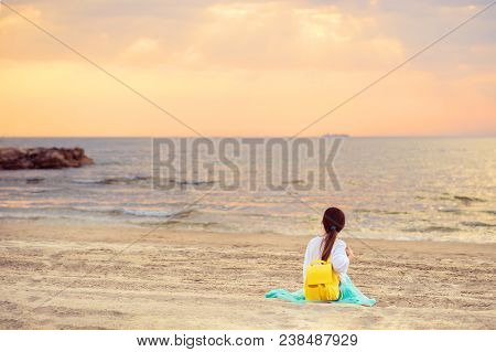 Young Woman With Yellow Backpack Sitting On Sandy Beach Looking At Sea And Sunset. Adriatic Sea In D