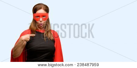 Middle age super hero woman wearing red mask and cape happy and surprised cheering expressing wow gesture, pointing with finger isolated blue background