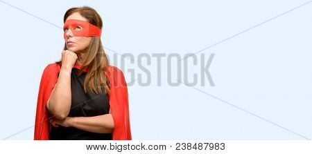 Middle age super hero woman wearing red mask and cape thinking and looking up expressing doubt and wonder isolated blue background