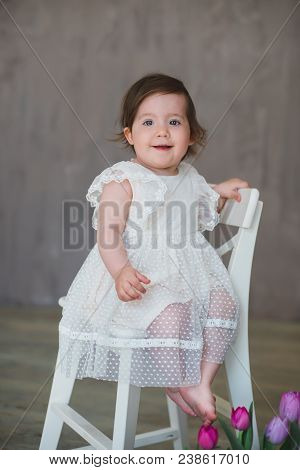 Beautiful Smiling Little Girl In White Lace Dress Among Tulips, Sitting On A White Chair.