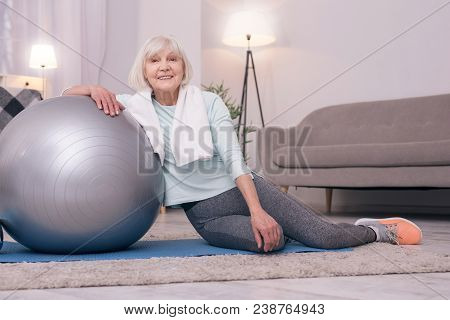 Pleasant Recreation. Charming Senior Woman Sitting On A Mat And Leaning On A Yoga Ball While Regaini