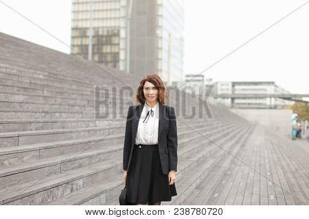 Happy Businesswoman Standing On Stairs With Bag And High Buildings In Background. Concept Of Profess