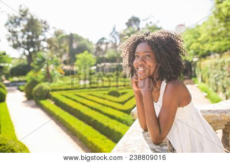 Dreamy Young Black Woman Leaning On Stone Railing In Park. Lady Standing With Beautiful Garden In Ba