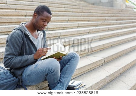 Young Serious African-american Man Reading Book, Sitting On Campus Stairs, Studying And Preparing Fo