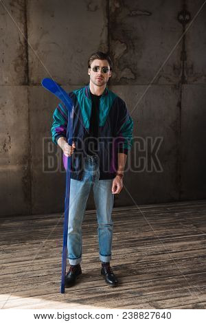 Stylish Young Man In Vintage Windcheater And Sunglasses With Hockey Stick Looking At Camera