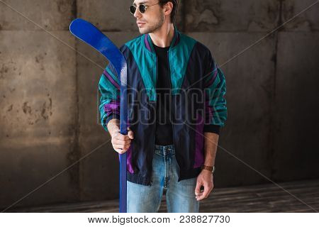 Stylish Young Man In Vintage Windcheater And Sunglasses With Hockey Stick Looking Away