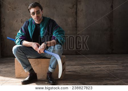 Handsome Young Man In Vintage Windcheater With Hockey Stick Sitting On Wooden Box