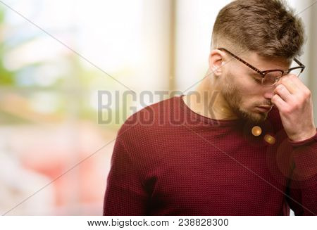 Handsome young man with sleepy expression, being overworked and tired, rubbes nose because of weariness