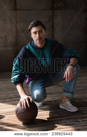 Stylish Young Man In Vintage Windcheater With Basketball Ball Sitting Squats On Wooden Floor